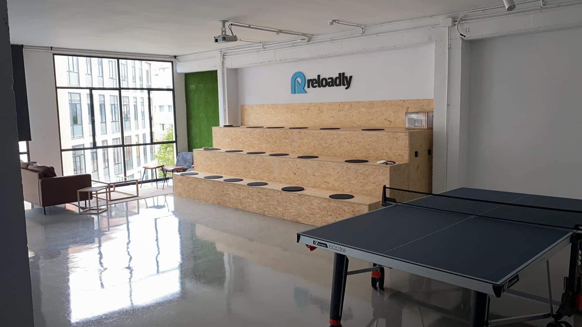 Zona de encuentro de las oficinas, con la grada de tablero OSB bajo el rótulo de Reloadly y la mesa de ping-pong