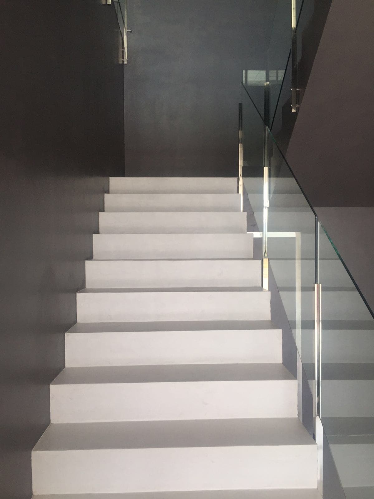 La escalera revestida de microcemento, con los peldaños claros, las paredes grises y la barandilla de vidrio