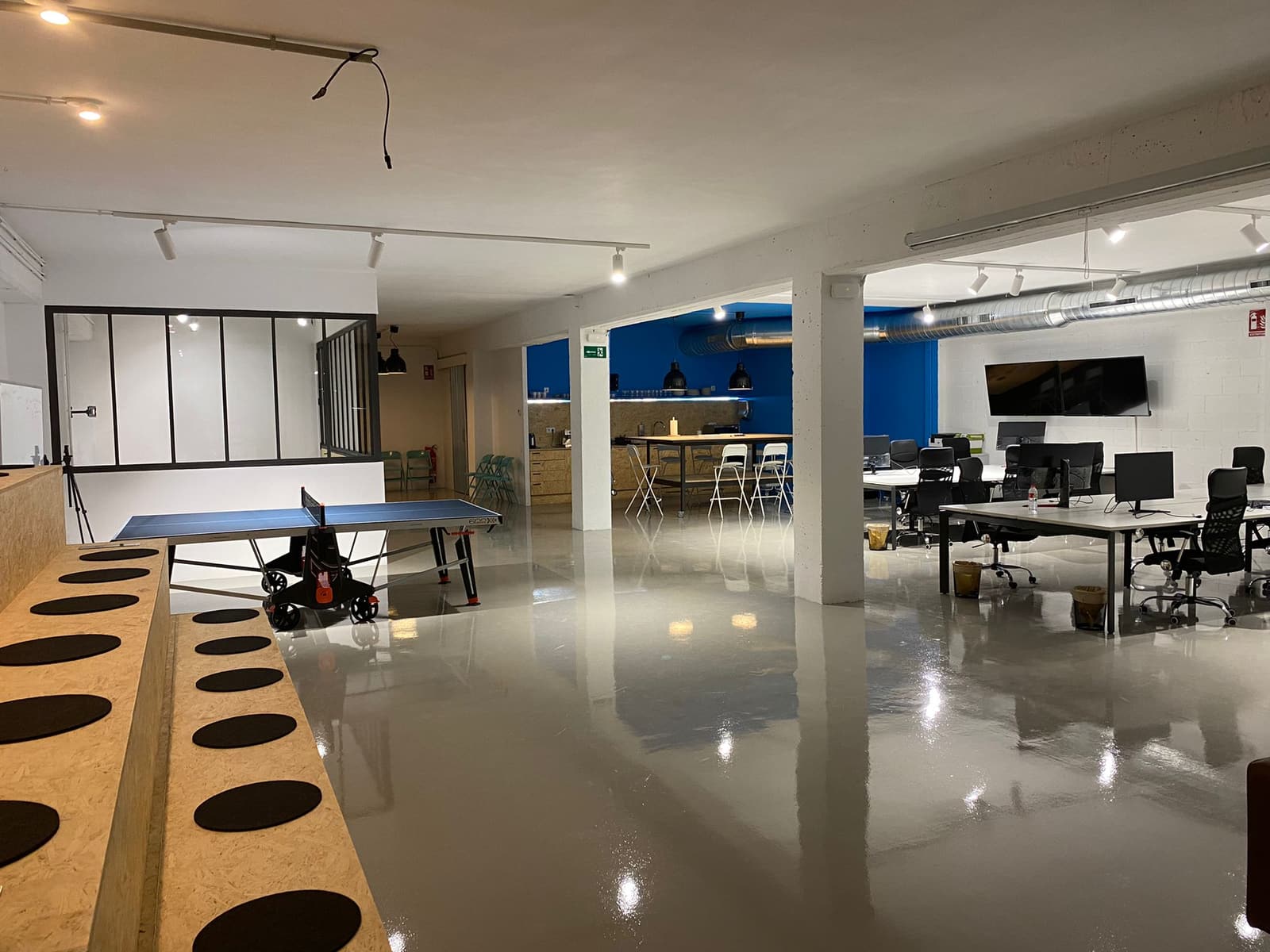 Vista general del espacio diáfano: la grada, la mesa de ping-pong, la cocina al fondo y los puestos de trabajo a la derecha