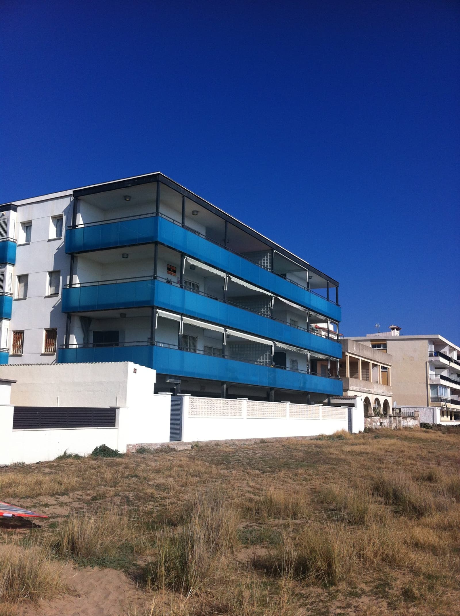 El edificio en esquina visto desde la duna, con las bandas de balcones azules apiladas en tres plantas