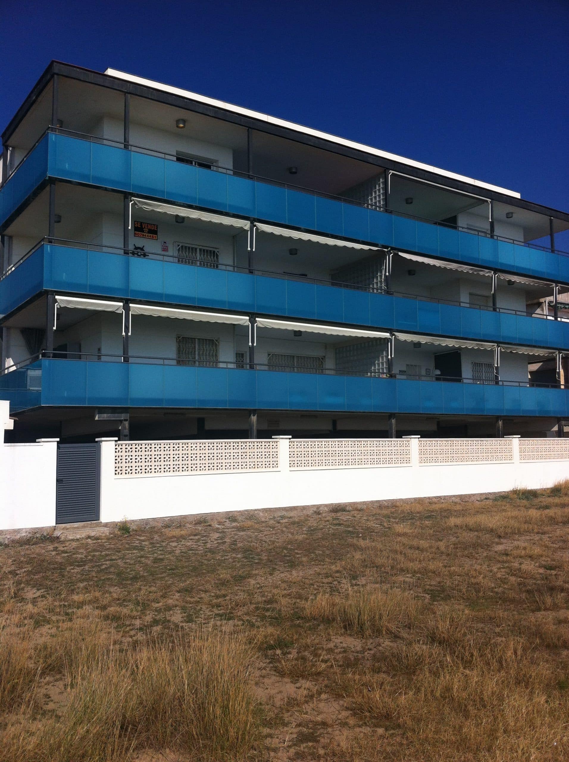 Fachada del edificio frente a la playa de Castelldefels, con tres plantas de balcones de vidrio azul y toldos, vista desde las dunas